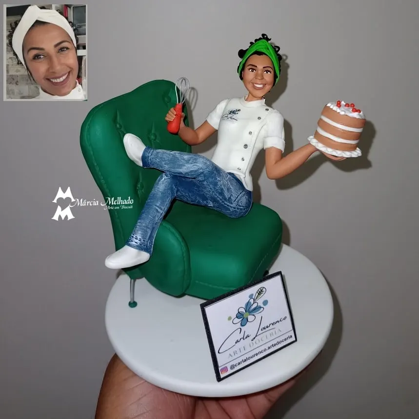 Boneca confeiteira em poltrona com bolo e batedor Confeiteira de biscuit de dólmã branca e turbante verde sentada em poltrona verde segurando bolo e batedor de arame