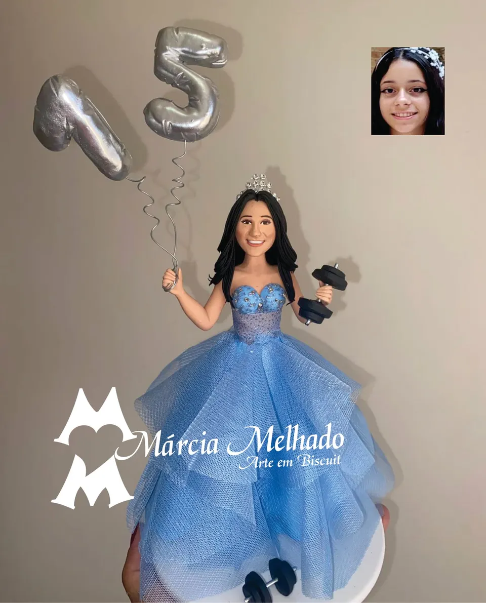 Boneca de 15 anos com vestido azul, tiara e halteres Boneca de biscuit de debutante com tiara e vestido azul de tule segurando balões do número 15 e um halter