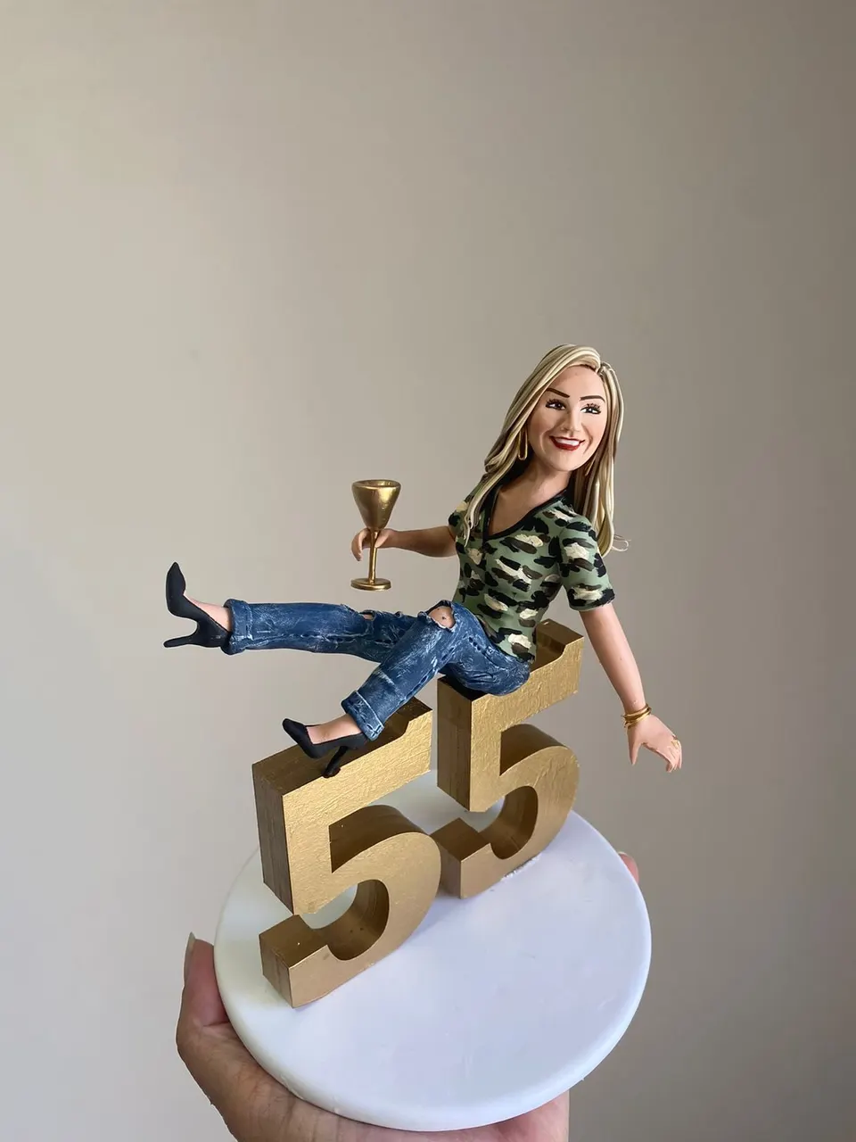 Boneca de aniversário 55 anos sentada no número com taça Boneca de biscuit loira sentada sobre o número 55 dourado, com calça jeans, salto alto e taça dourada na mão
