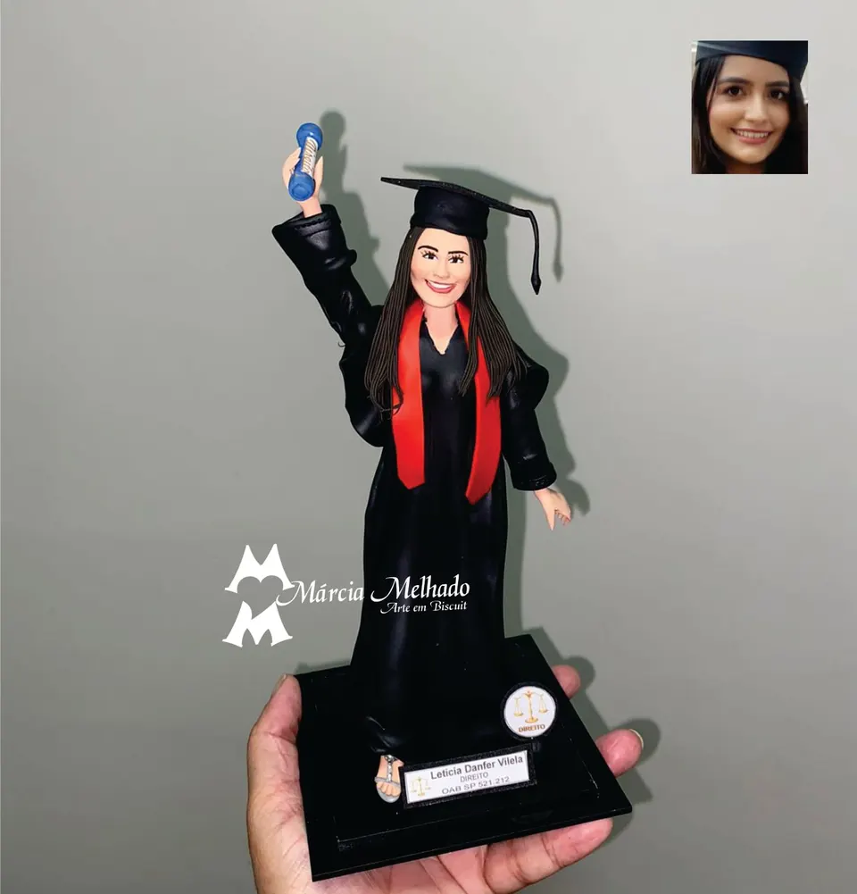 Boneca de formatura de Direito com diploma e capelo Boneca de biscuit de formanda de Direito com beca preta, faixa vermelha e capelo, erguendo o diploma azul em uma base preta
