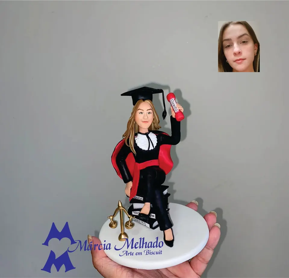 Boneca de formatura em Direito sentada sobre livros com balança Boneca de biscuit de formanda em Direito com beca, capelo e canudo, sentada sobre livros ao lado de balança dourada