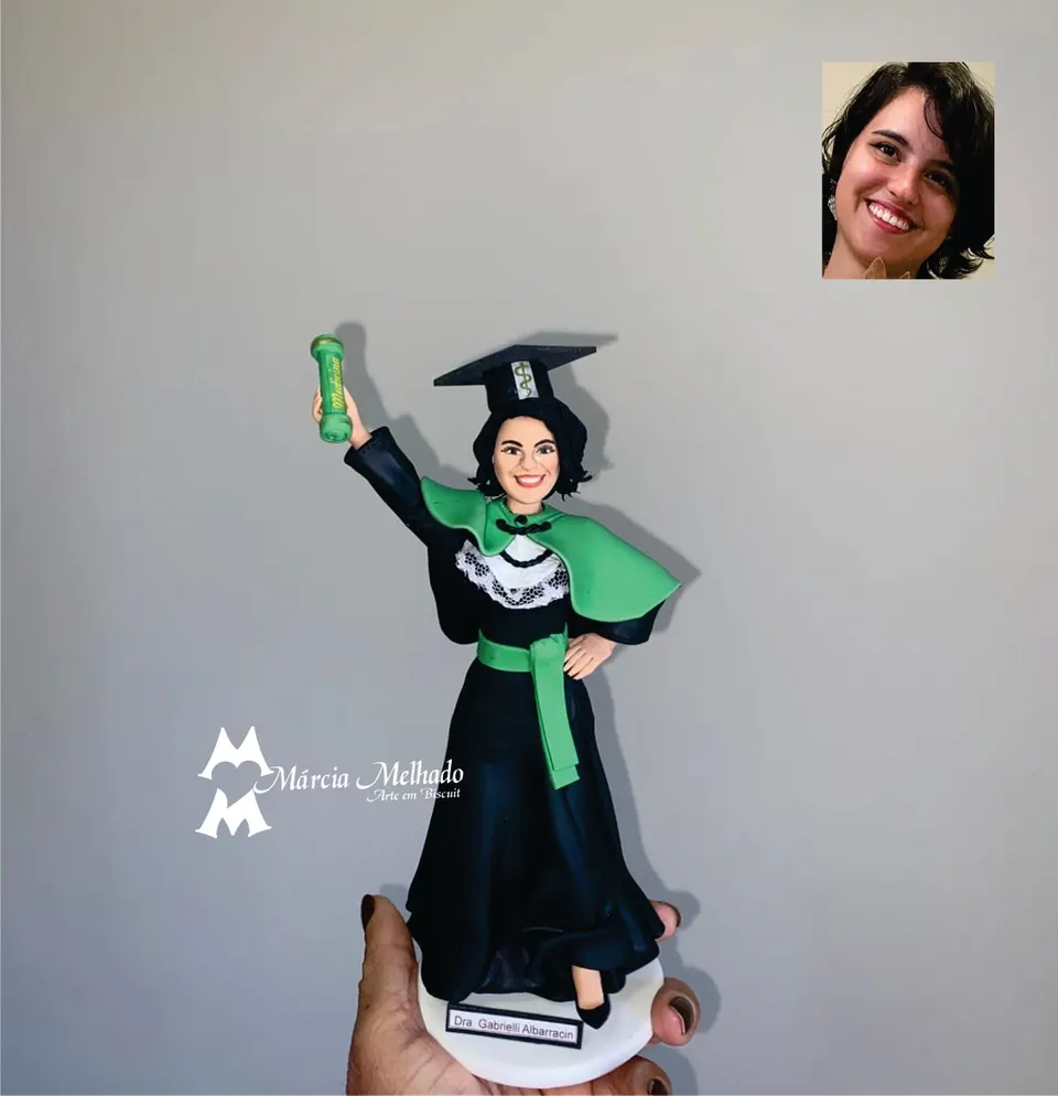 Boneca de formatura em Medicina com capelo e canudo verde erguido Boneca de biscuit de formanda de Medicina com beca preta, capa e faixa verdes e capelo, erguendo o canudo verde