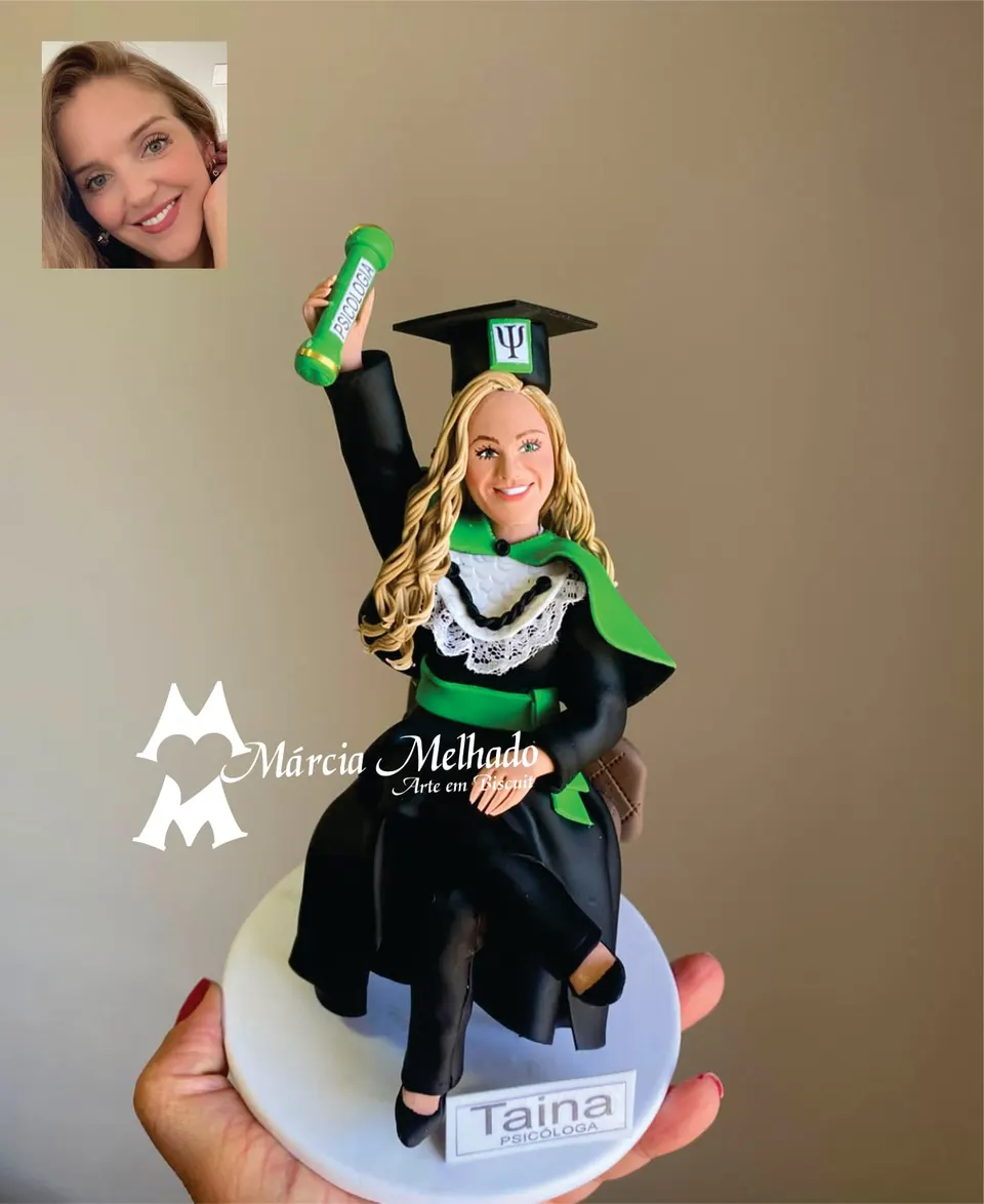 Boneca de formatura em psicologia sentada com canudo erguido Boneca de biscuit de formanda em psicologia sentada, com beca preta, faixa verde, capelo e canudo erguido