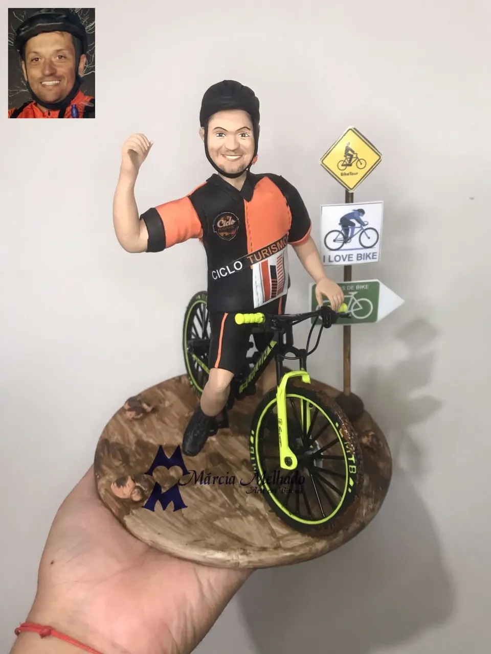 Boneco ciclista pedalando mountain bike com placas de trilha Boneco de biscuit ciclista de capacete pedalando mountain bike preta e verde com punho erguido ao lado de placas de trilha