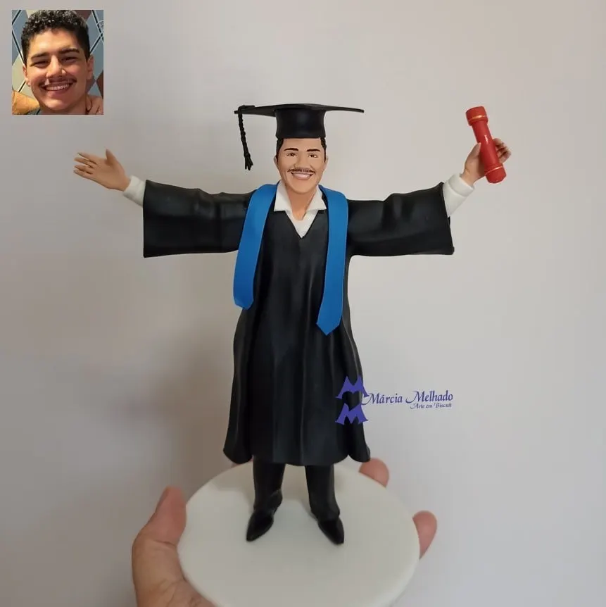 Boneco de formatura com beca, capelo e canudo Boneco de biscuit formando de beca preta, faixa azul-clara e capelo, de braços abertos segurando canudo vermelho