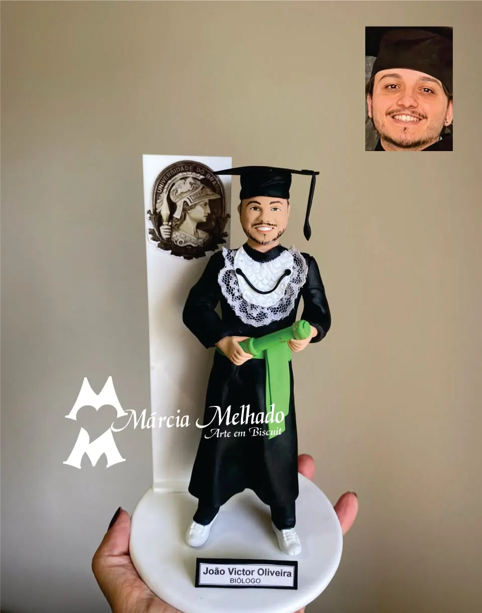 Boneco de formatura em Biologia com canudo verde e brasão da faculdade Boneco de biscuit de formando com beca preta, capelo e canudo verde, diante de painel branco com brasão da universidade