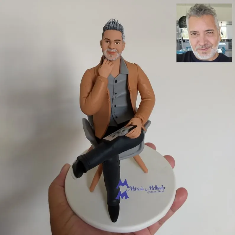 Boneco de homem grisalho sentado em poltrona com tablet Boneco de biscuit de homem grisalho com jaqueta caramelo sentado de pernas cruzadas em poltrona cinza segurando um tablet
