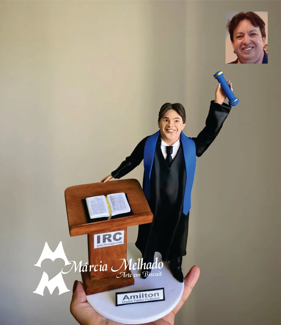Boneco de líder religioso no púlpito com Bíblia e diploma Boneco de biscuit de pregador com beca preta e faixa azul erguendo diploma ao lado de púlpito de madeira com Bíblia aberta