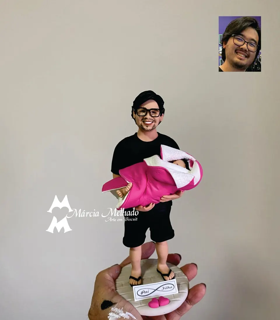 Boneco de pai com bebê no colo e plaquinha pai e filha Boneco de biscuit de homem de óculos e camiseta preta segurando bebê em manta rosa, com plaquinha pai e filha