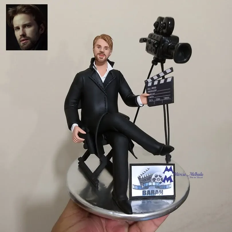 Boneco diretor de cinema com câmera e claquete Boneco de biscuit de terno preto sentado em cadeira de diretor segurando claquete, com câmera de cinema em tripé ao lado