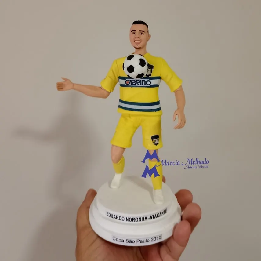 Boneco jogador de futebol com uniforme amarelo e bola no peito Boneco de biscuit de jogador de futebol com uniforme amarelo dominando a bola no peito, sobre base com plaquinhas