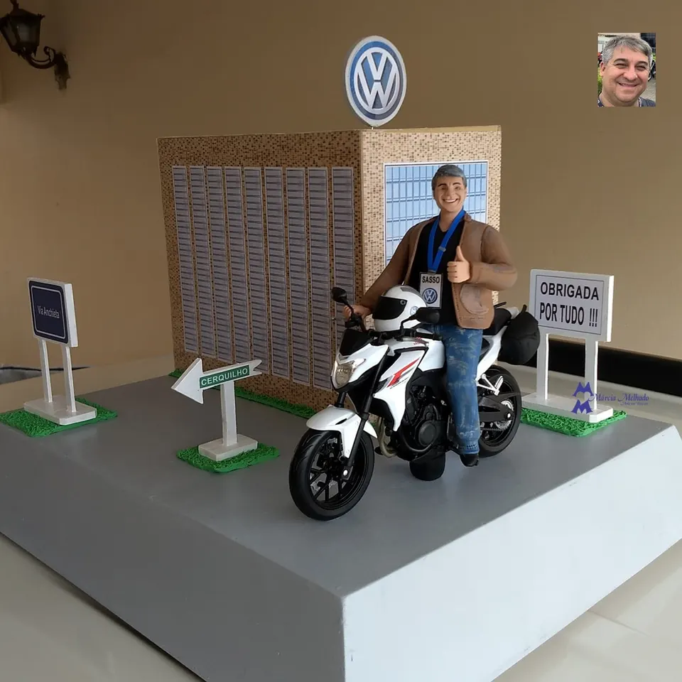 Boneco motociclista com moto e prédio da Volkswagen Boneco de biscuit de homem grisalho com jaqueta de couro em moto branca diante de prédio da Volkswagen e placas de rua
