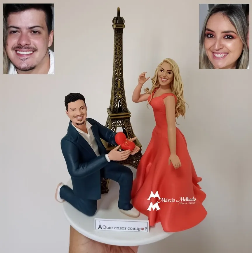 Bonecos de casal em pedido de casamento com Torre Eiffel Boneco de terno azul ajoelhado com caixinha de coração e aliança diante de boneca loira de vestido vermelho e Torre Eiffel