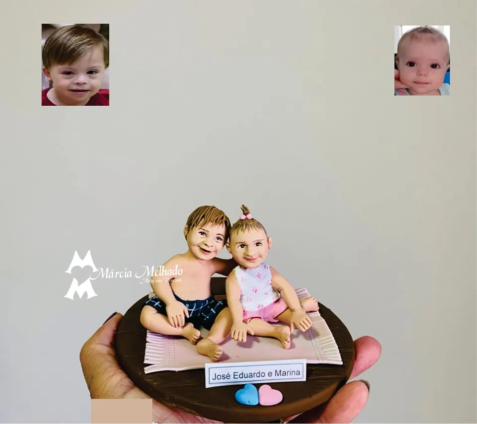 Bonecos de irmãos pequenos sentados em tapete com corações Bonecos de biscuit de dois irmãos pequenos sentados num tapete rosa sobre base marrom, com plaquinha e corações azul e rosa