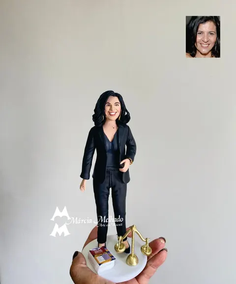 Boneca de biscuit de mulher de terninho preto ao lado de balança da justiça dourada e livro Vade Mecum em base branca