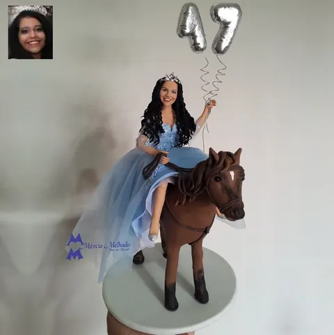 Debutante de biscuit com vestido azul e tiara montada em cavalo marrom segurando balões prateados do número 15