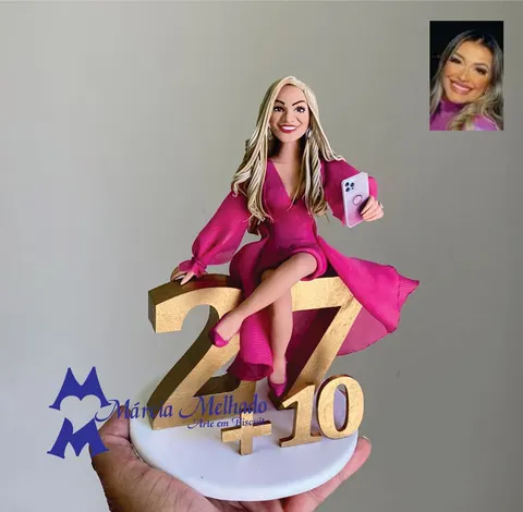 Boneca de biscuit loira com vestido pink de mangas bufantes sentada sobre os números dourados 27+10, segurando um celular