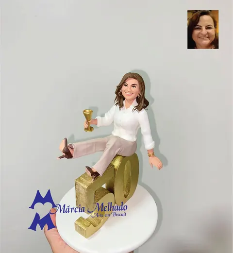 Boneca de biscuit de mulher de camisa branca e calça bege sentada sobre o número 50 dourado, segurando uma taça dourada