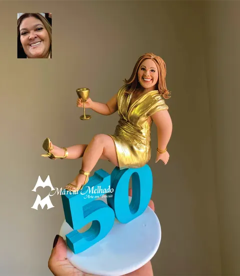 Boneca de biscuit de vestido dourado e sandálias douradas, com taça na mão, sentada sobre o número 50 azul turquesa