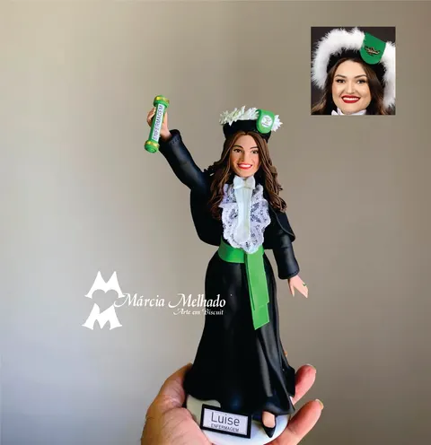 Boneca de biscuit de formanda de Enfermagem com beca preta, jabô de renda, faixa verde e capelo florido, erguendo o diploma