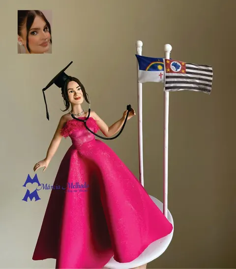 Boneca de biscuit formanda com vestido pink, capelo e estetoscópio ao lado das bandeiras de Pernambuco e São Paulo