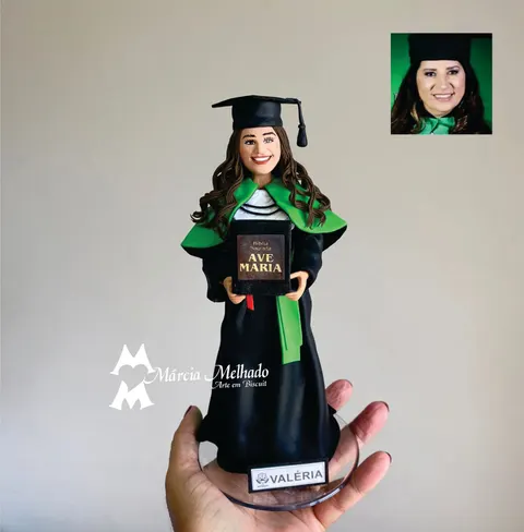 Boneca de biscuit de formanda de Nutrição com beca preta, capa verde e capelo, segurando uma Bíblia Sagrada com as duas mãos