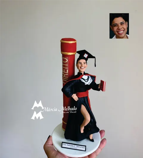 Boneca de biscuit formanda em Direito de beca preta com faixa vermelha, capelo e livro, ao lado de canudo gigante vermelho
