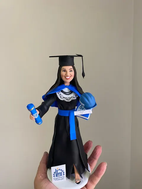 Boneca de biscuit formanda de beca preta e faixa azul segurando diploma, livros e capacete de engenheira