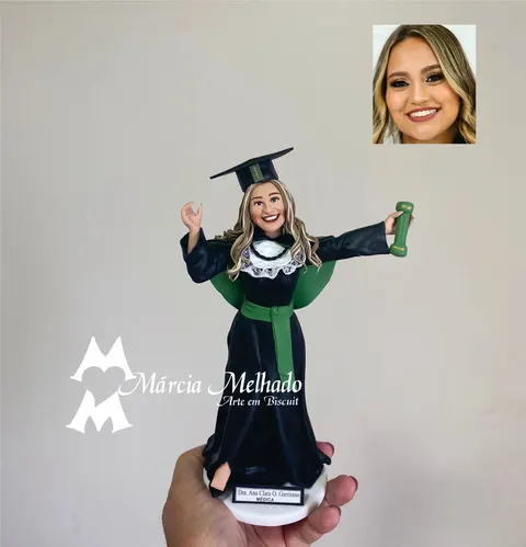 Boneca de biscuit de formanda loira em medicina com beca preta, faixa verde, capelo e canudo nas mãos