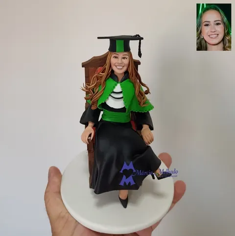 Boneca de biscuit de formanda com beca preta, capa e faixa verdes e capelo, sentada em cadeira de madeira de pernas cruzadas