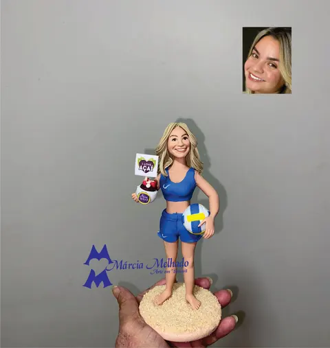 Boneca de biscuit loira com uniforme azul de vôlei de praia segurando bola e pote de açaí sobre base de areia