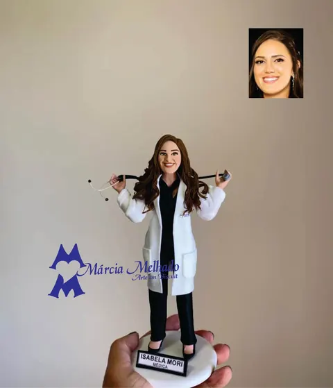 Boneca de biscuit de médica com jaleco branco, roupa preta e estetoscópio apoiado nos ombros, em base branca com plaquinha