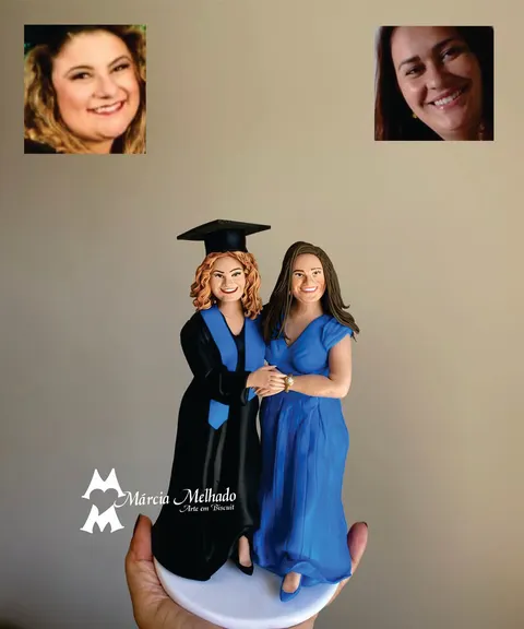 Duas bonecas de biscuit de mãos dadas: formanda de beca preta com faixa azul e capelo ao lado de mulher de vestido azul longo
