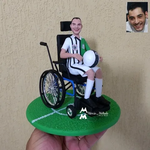 Boneco de biscuit em cadeira de rodas com camisa do Corinthians e do Palmeiras segurando bola de futebol sobre base de grama