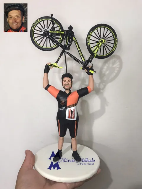 Boneco de biscuit ciclista de capacete e camisa laranja e preta erguendo uma mountain bike preta e verde acima da cabeça