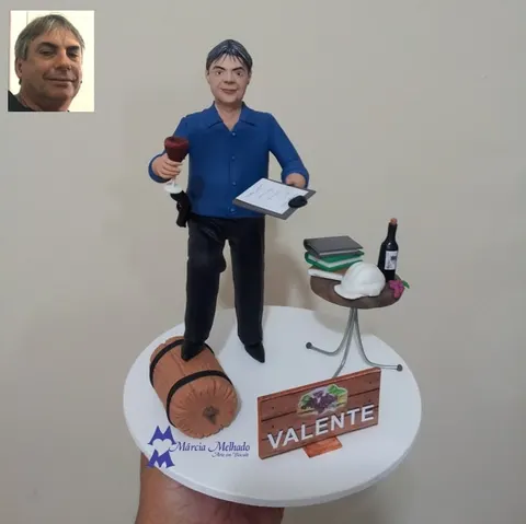 Boneco de biscuit de homem grisalho com taça de vinho e prancheta, ao lado de barril, mesa com livros e capacete branco