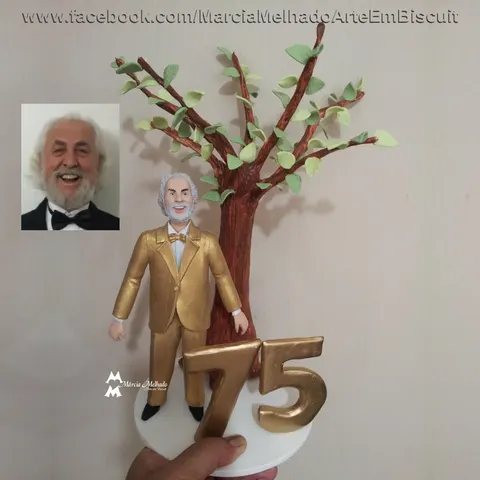 Boneco de biscuit de senhor de barba branca com terno dourado em frente a uma árvore, com o número 75 em dourado na base