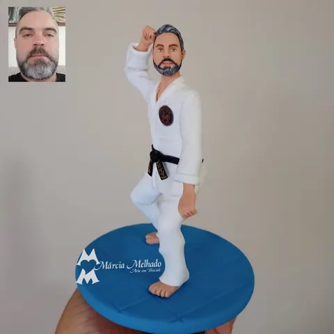 Boneco de biscuit de homem barbudo com kimono branco e faixa preta em pose de defesa, descalço sobre base azul de tatame