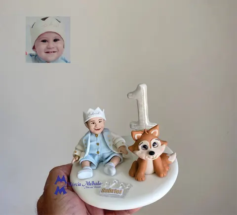 Bebê de biscuit sentado com coroa e roupa de príncipe azul-clara ao lado de uma raposinha e de um número 1 perolado