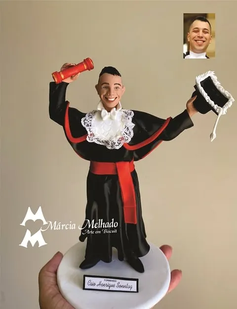 Boneco de biscuit formando de beca preta com faixa vermelha, erguendo canudo vermelho e capelo com pelúcia branca
