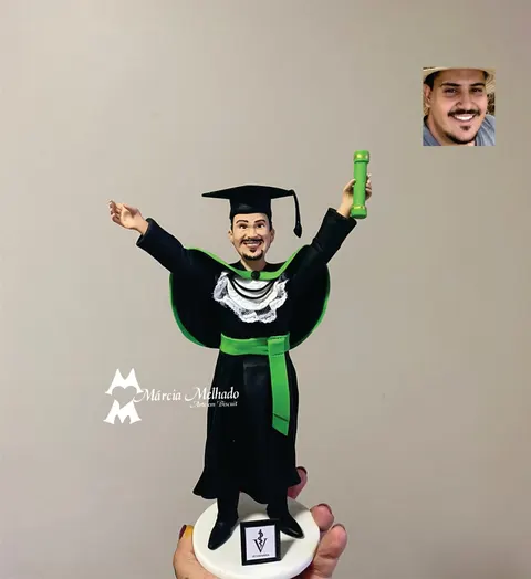 Boneco de biscuit de formando de veterinária com beca preta, faixa verde e capelo, braços erguidos com o canudo do diploma