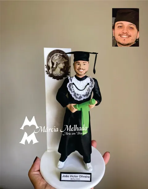 Boneco de biscuit de formando com beca preta, capelo e canudo verde, diante de painel branco com brasão da universidade