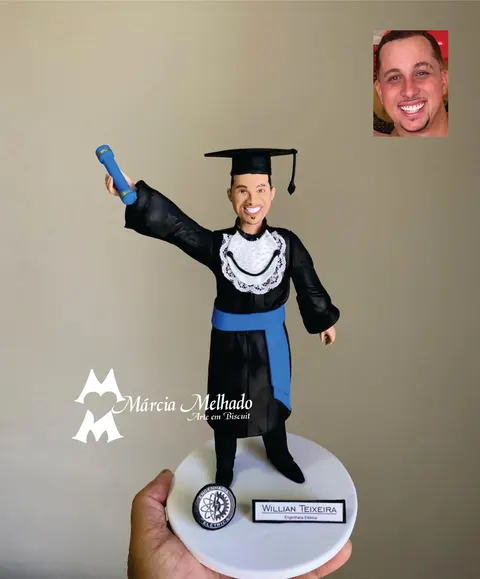 Boneco de biscuit de formando de engenharia elétrica com beca preta, faixa azul e capelo erguendo o canudo azul