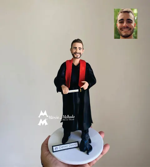 Boneco de biscuit formando com beca preta e faixa vermelha segurando diploma, com gato preto na base branca