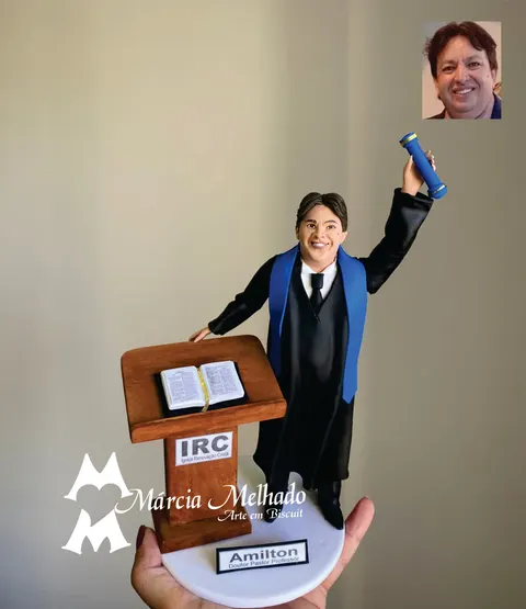 Boneco de biscuit de pregador com beca preta e faixa azul erguendo diploma ao lado de púlpito de madeira com Bíblia aberta