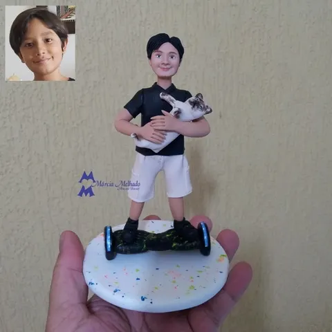 Boneco de biscuit de menino de polo preta e bermuda branca em cima de um hoverboard, com um gato branco e marrom no colo