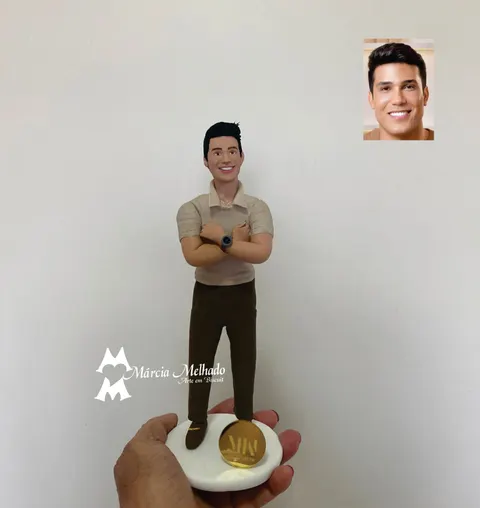 Boneco de biscuit de homem sorridente com polo bege, calça marrom e braços cruzados, sobre base branca com placa dourada