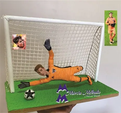 Boneco de biscuit de goleiro com uniforme laranja voando para defender a bola em um gol com rede, sobre base de grama.