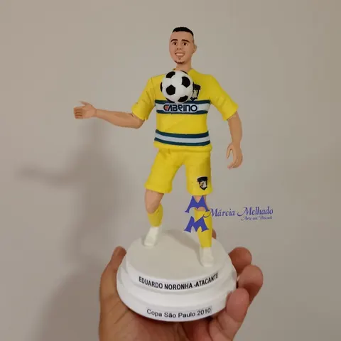 Boneco de biscuit de jogador de futebol com uniforme amarelo dominando a bola no peito, sobre base com plaquinhas