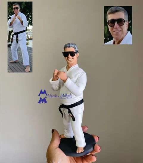 Boneco de biscuit de homem grisalho de óculos escuros com kimono branco e faixa preta em posição de luta sobre base preta.
